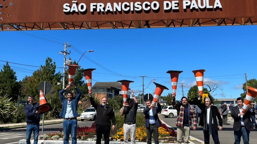 Oito homens lado a lado levantando cones de trânsito para o alto, abaixo do pórtico da cidade onde se lê São Francisco de Paula