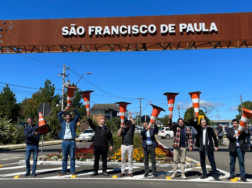 Oito homens lado a lado levantando cones de trânsito para o alto, abaixo do pórtico da cidade onde se lê São Francisco de Paula