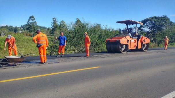 Uma máquina para prensar asfalto é vigiada por trabalhadores de uniforme laranja em um trecho da rodovia