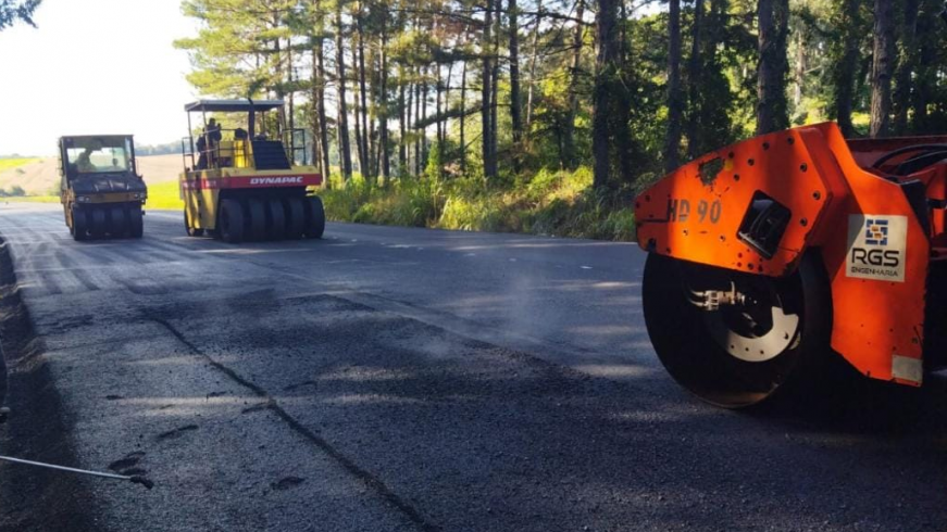 Três máquinas estão prensando o asfalto novo na rodovia. Ao fundo, pinheiros contrastam com o preto do asfalto e o laranja de uma das máquinas. Dá para ver a fumaça do asfalto quente.