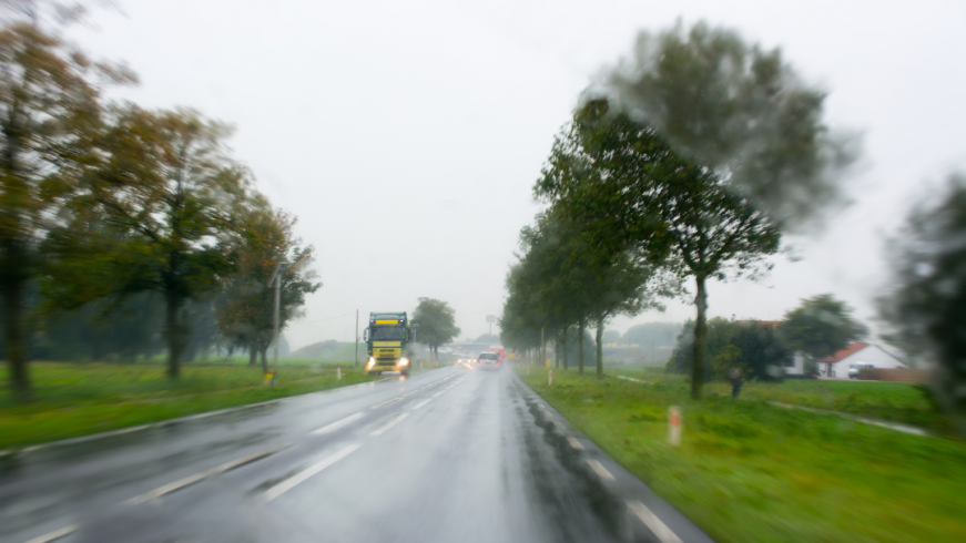 A foto retrata uma estrada molhada. O tempo está chuvoso, com pouca visibilidade. Ao fundo, um caminhão parado no acostamento e carros com farol ligado.